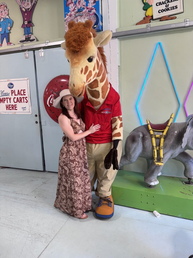 woman in brown dress with cream hat standing next to stuffed giraffe